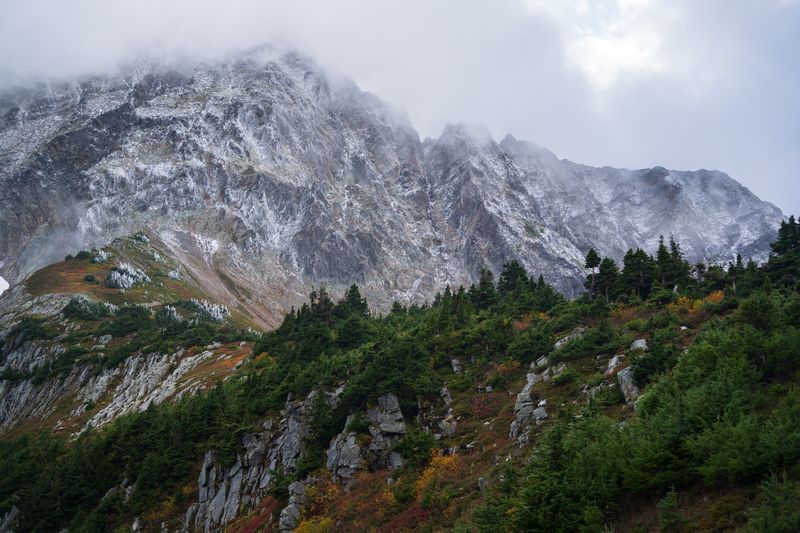 Cloud clinging to a jagged, snow-dusted ridge over a forested autumn hillside in the North Cascades